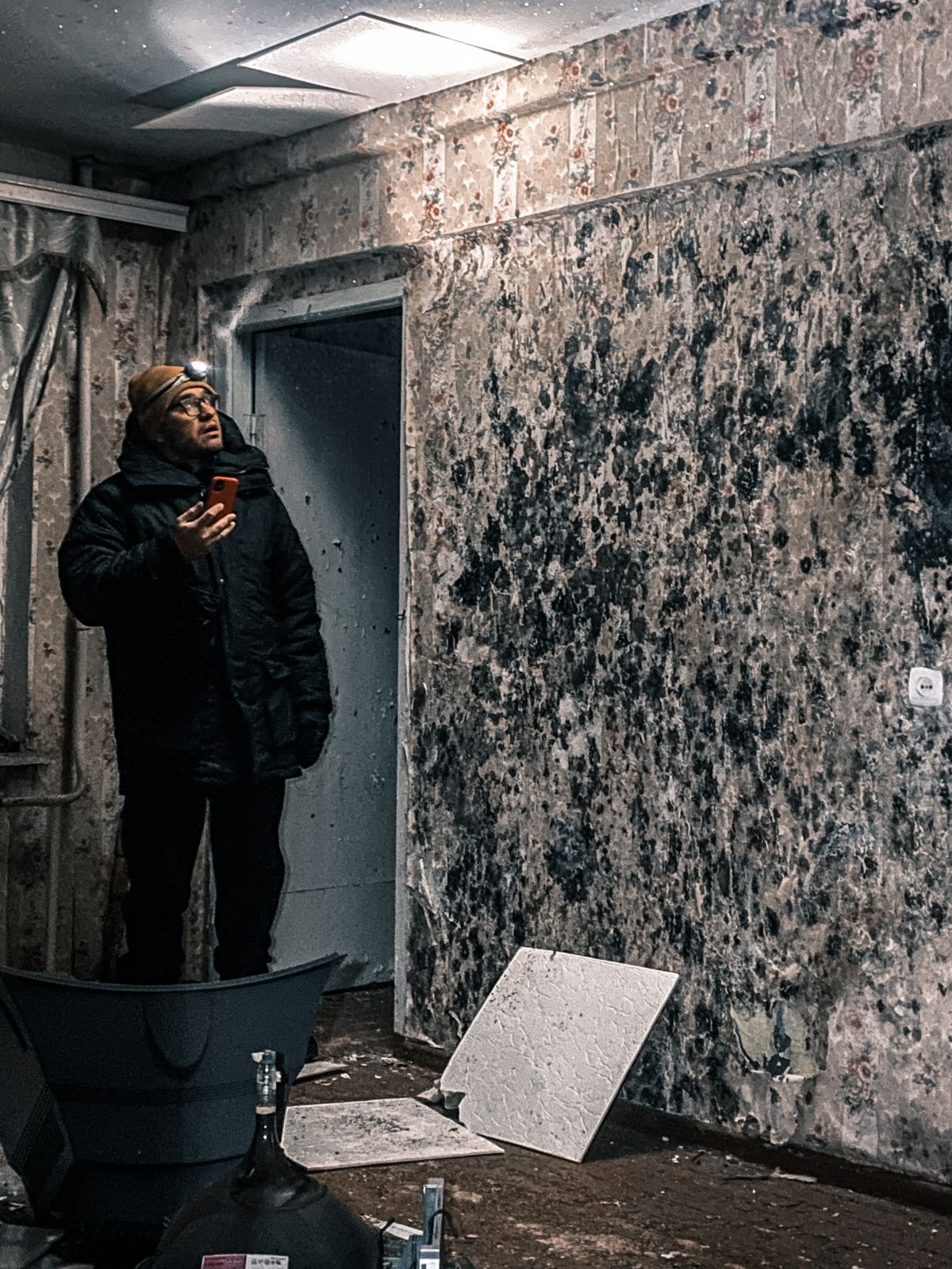Person wearing a headlamp photographing the damaged interior of a deteriorated older building with fallen ceiling tiles on the floor
