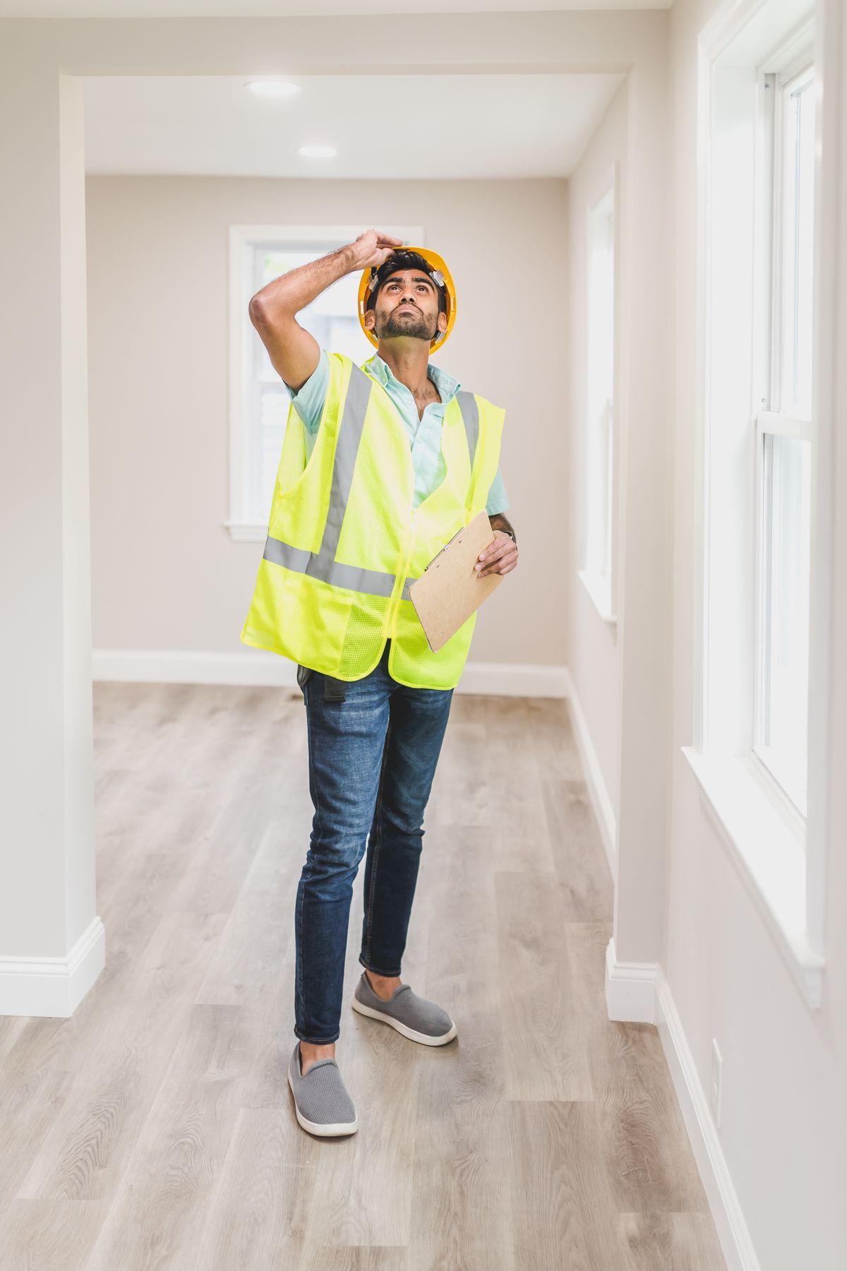 Inspector in a hard hat and safety vest standing in an empty room, looking up toward the ceiling with a clipboard in hand