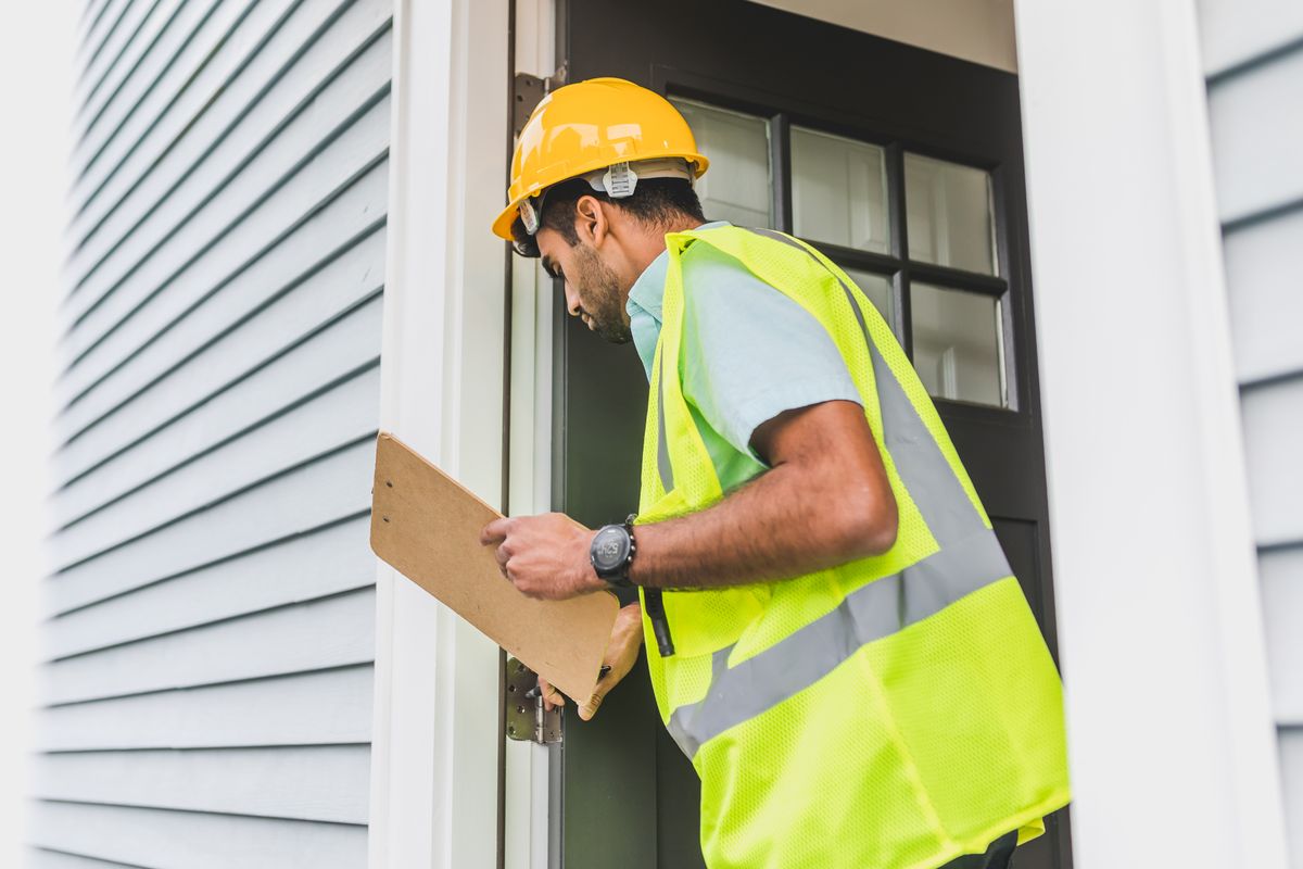 Inspector in a hard hat and reflective vest examining a building's exterior door frame while holding a clipboard